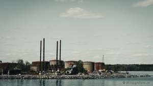 Verlassene Industrieanlage auf einer Insel, rostige Öltanks und vier Schornsteine vor bewölktem Himmel, umgeben von Wasser