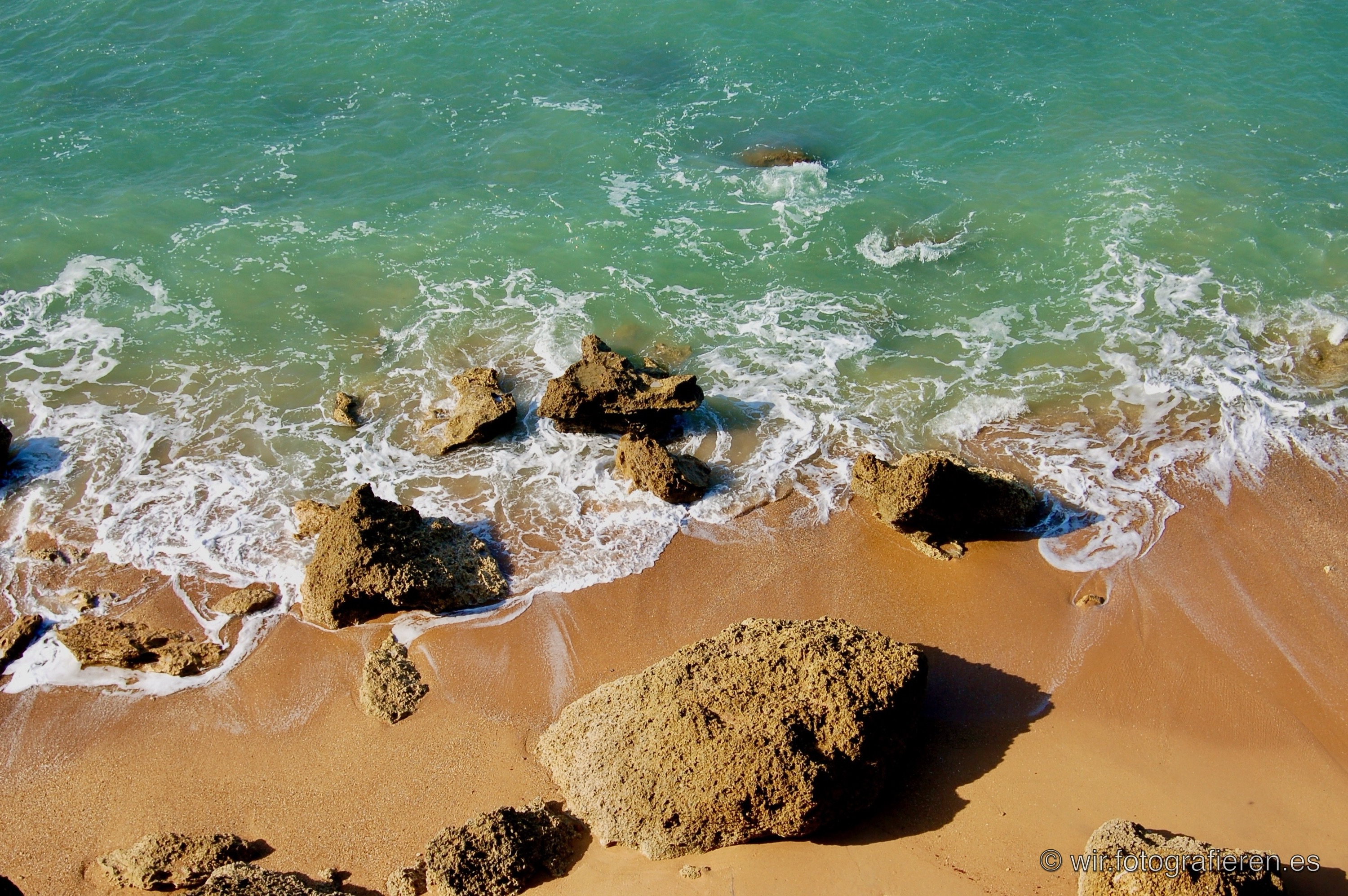 Vogelperspektive auf Atlantikstrand der Algarve, Portugal, türkisfarbenes Wasser trifft auf warmen orangefarbenen Sand, Felsen im Brandungsbereich, lebhafte Farbkontraste