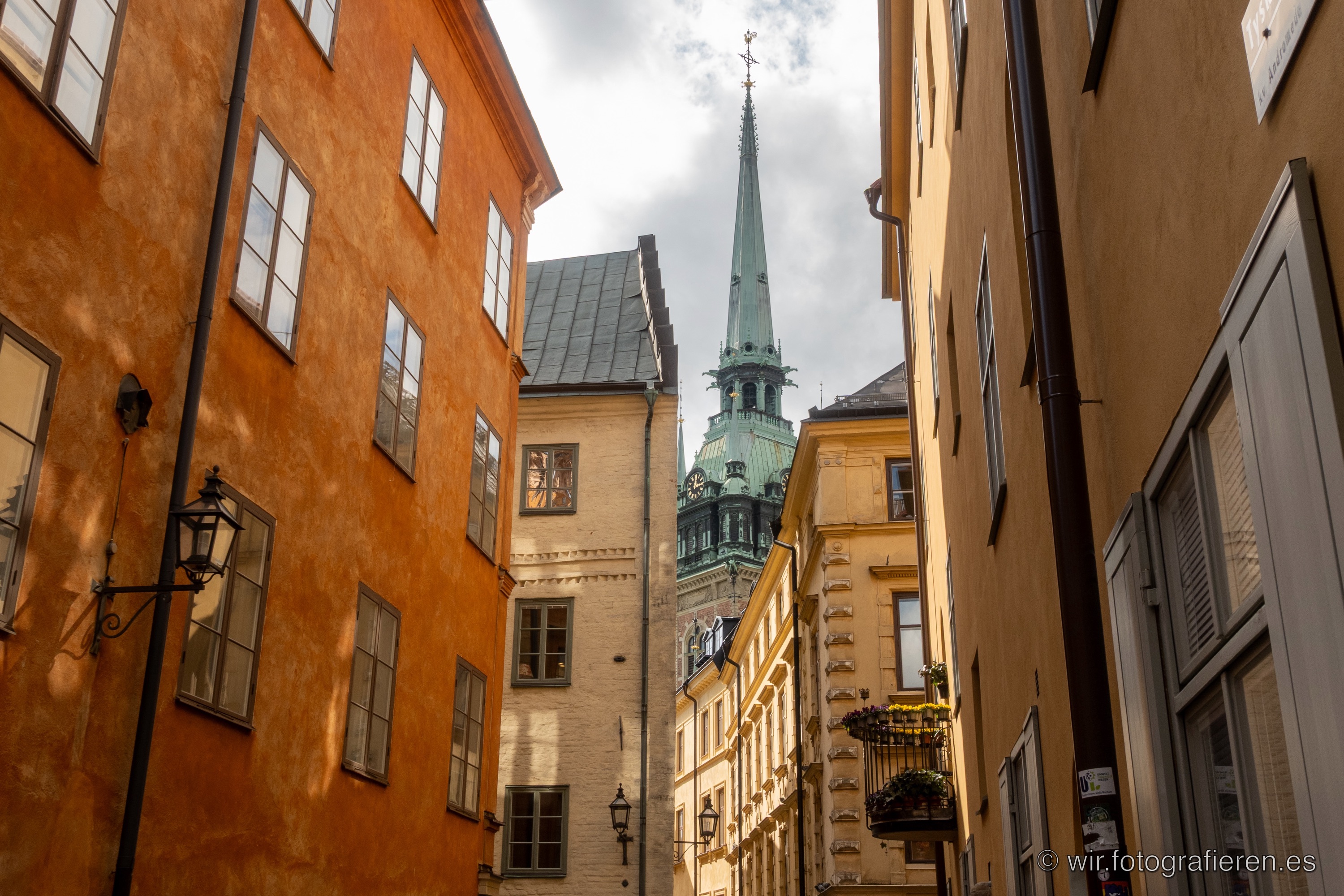 Enge Gasse in Stockholms Gamla Stan mit warmen Ockerfassaden, grüner Kupferspitzturm der Deutschen Kirche zwischen den Häusern, südlich wirkendes Licht